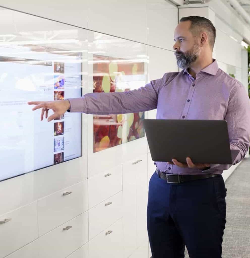 A man wearing a light pink button-up shirt and dark pants is standing in an office while holding a laptop. He is pointing towards a large screen on the wall displaying various images and text. The office space is modern with white cabinetry and bright lighting.