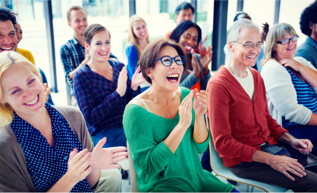 A diverse group of people, both men and women, are sitting in chairs and clapping while smiling and laughing. They are in a bright room with large windows. Some individuals are wearing glasses, and most are dressed casually. The atmosphere appears cheerful and lively, enhanced by discussions on AI-enabled outreach.