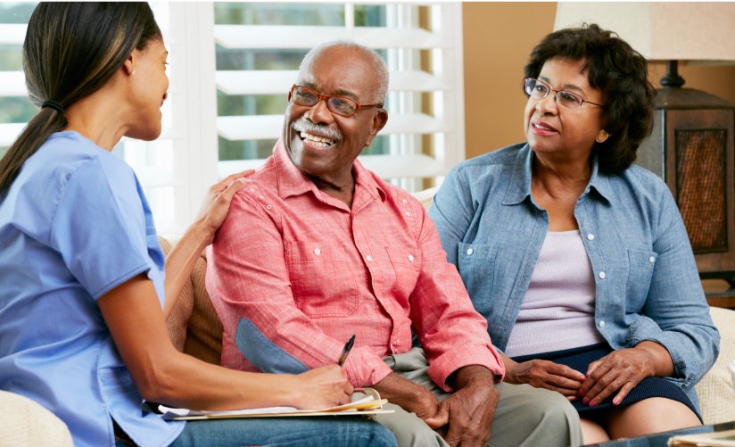 A young nurse wearing a blue uniform sits on a couch, smiling and talking with an elderly couple. The elderly man, wearing glasses and a red shirt, smiles back while the elderly woman in a blue shirt looks on attentively. The setting appears to be a cozy living room.