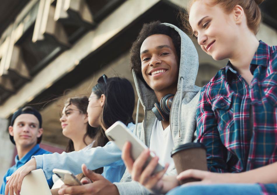 A group of five young people sit together outdoors. They are casually dressed, some with hats and one with headphones around his neck. They are all smiling, holding smartphones and drinks, enjoying each other's company under a structure or building.