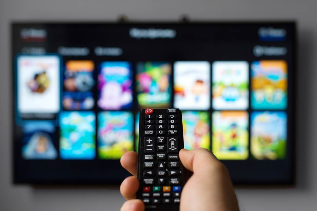 A hand holding a remote control is pointed at a TV screen. The TV displays several colorful, blurred icons of various streaming service apps. The remote is in sharp focus while the TV screen remains slightly out of focus in the background.