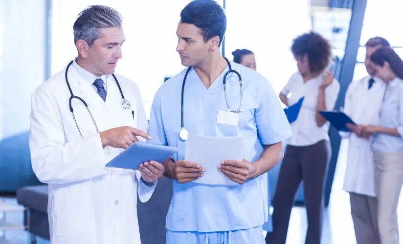 Two healthcare professionals, one in a white coat and the other in blue scrubs, discuss documents in a modern medical office. Both hold paperwork and stethoscopes are visible around their necks. Other medical staff are seen working in the background.