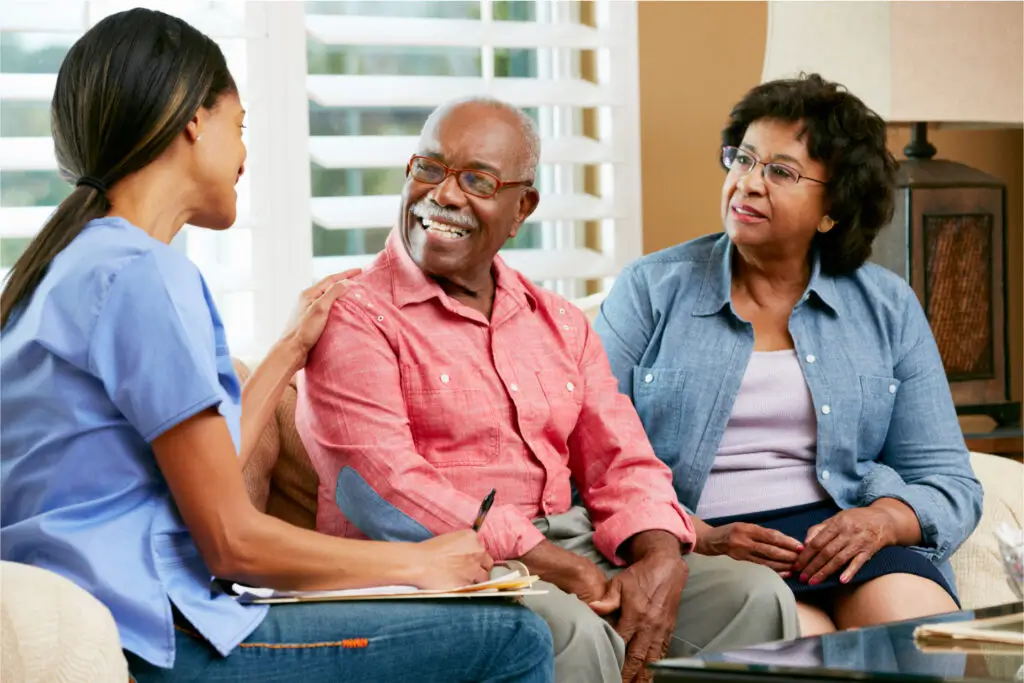 A healthcare worker in a blue uniform sits and engages in conversation with an older couple. The man and woman are sitting close together on a couch, smiling and looking at the healthcare worker. The background includes window blinds and a lamp.