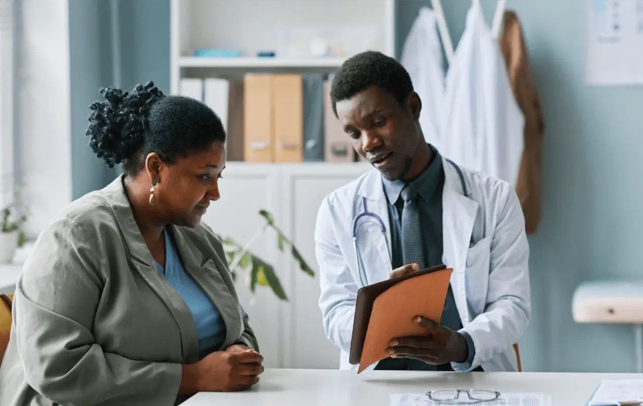 A healthcare professional wearing a white coat and stethoscope is showing a tablet to a seated patient in a medical office, emphasizing comprehensive patient care. The patient appears to be attentively listening. The office has folders, plants, and medical gowns hanging in the background.